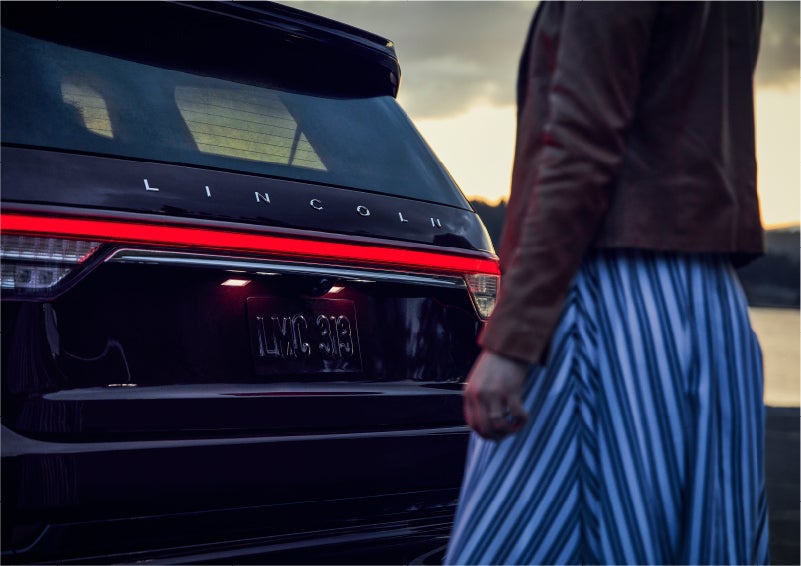 A person is shown near the rear of a 2023 Lincoln Aviator® SUV as the Lincoln Embrace illuminates the rear lights | West Point Lincoln in Houston TX